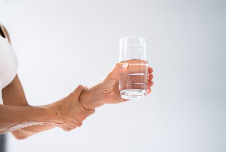 Dystonie, définition, cervicale, cause, prise en charge. Image d'une femme atteinte de dystonie. Elle tient fermement son bras gauche au bout duquel sa main porte un verre rempli d'eau.