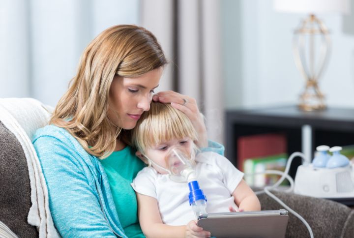 Mucoviscidose : symptômes, espérance de vie, traitement. Image d'une maman enfant sur les genoux pendant qu'il aspire un traitement respiratoire avec un masque sur la bouche et le nez. 