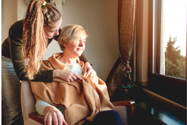 Journée mondiale des soins palliatifs 2024 : une jeune femme installe un châle autour d'une dame assise regardant au travers de la fenêtre.